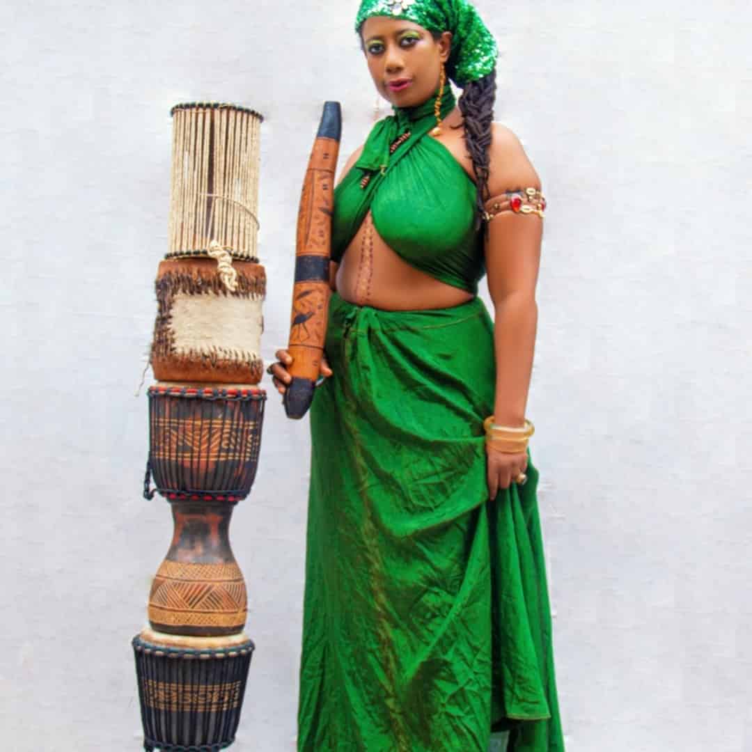 ElnanaAfrika in green traditional attire holding a talking drum stick beside a stack of African drums
